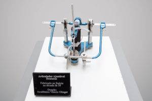 Um articulador ajustável dentário modelo Dentatus fabricado na Suécia, na década de 1950. A ferramenta é composta por uma estrutura metálica azul e prateada, com várias partes móveis e ajustáveis, usada para simular movimentos mandibulares. Ao lado, uma placa preta com texto branco fornece informações: "Articulador Ajustável Dentatus, Fabricado na Suécia na década de 50, Doação: Acadêmico Vinicio Olinger". A ferramenta está disposta sobre uma superfície branca.