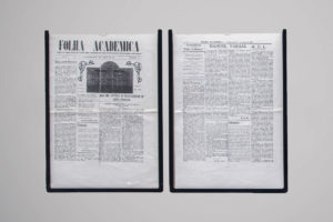 Fotografia de duas páginas de um jornal antigo emolduradas lado a lado. O título na página da esquerda é "Folha Acadêmica", escrito em letras grandes e estilizadas no topo. Ambas as páginas estão preenchidas com colunas de texto denso, típico de jornais, e há uma fotografia central de um prédio na página da esquerda. A imagem sugere um estilo de jornal impresso antigo, com layout tradicional e letras pequenas.