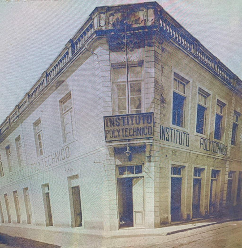 Prédio de esquina com fachada clara e detalhes em azulejos, com janelas retangulares e portas no nível térreo. Na fachada, há várias inscrições "INSTITUTO POLYTECHNICO" em letras grandes, inclusive em uma placa no canto superior do edifício. O estilo arquitetônico é clássico, com parapeito ornamental no topo. A imagem é a reprodução de uma fotografia antiga, com tons amarelados.