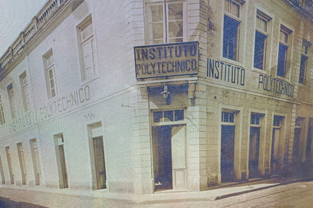 Prédio de esquina com fachada clara e detalhes em azulejos, com janelas retangulares e portas no nível térreo. Na fachada, há várias inscrições "INSTITUTO POLYTECHNICO" em letras grandes, inclusive em uma placa no canto superior do edifício. O estilo arquitetônico é clássico, com parapeito ornamental no topo. A imagem é a reprodução de uma fotografia antiga, com tons amarelados.