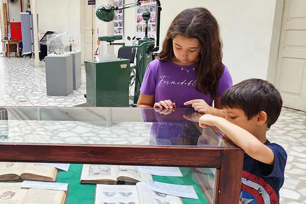 Uma menina e um menino observam atentamente livros e documentos expostos em uma vitrine de vidro em um ambiente de museu. A menina usa camiseta roxa e está com o dedo próximo à vitrine, enquanto o menino, com camiseta azul, também observa de perto. Ao fundo, há objetos museológicos, incluindo uma cadeira odontológica antiga e outros equipamentos.