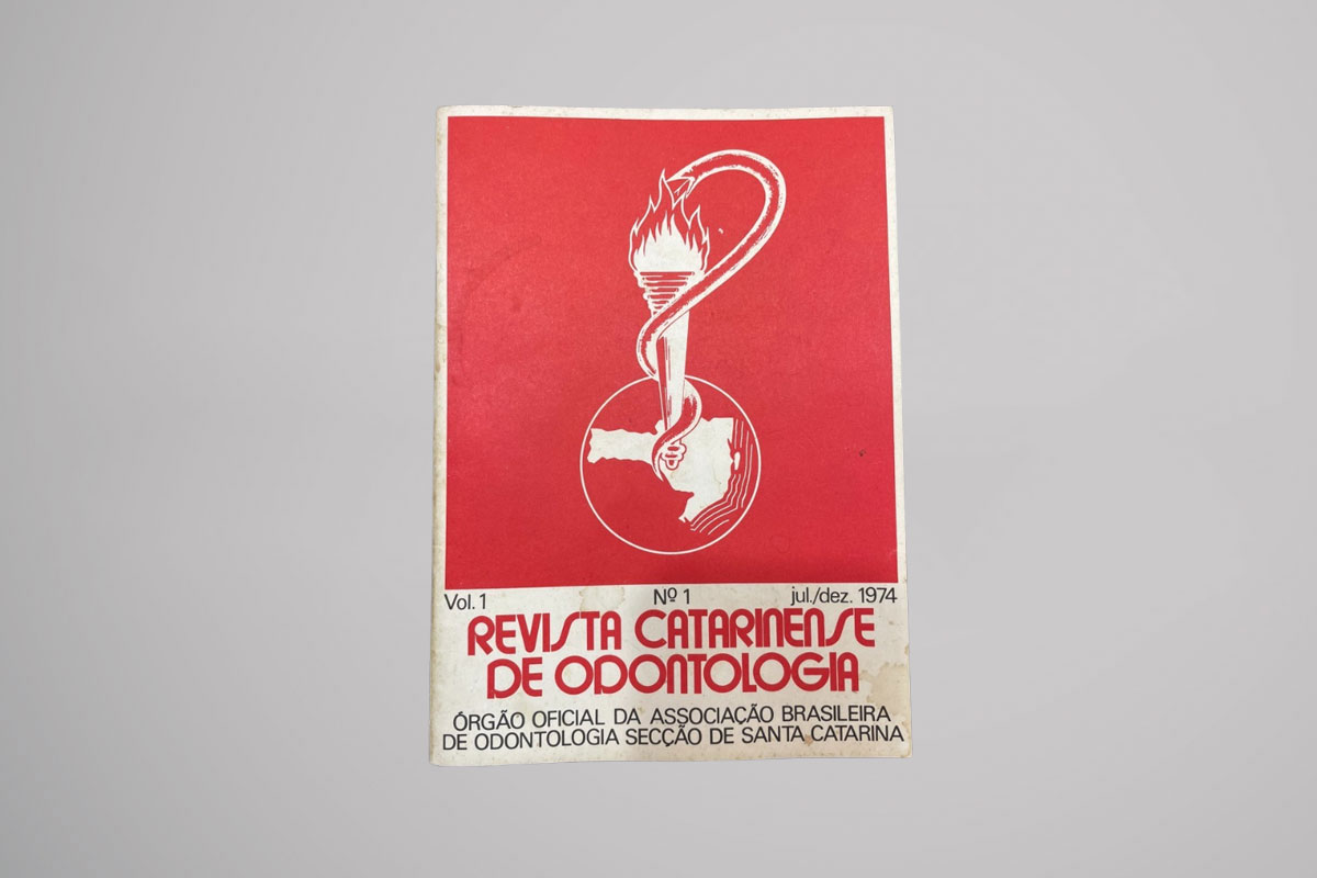 Capa da "Revista Catarinense de Odontologia", volume um, número um, referente ao período de julho a dezembro de mil novecentos e setenta e quatro. O fundo da capa é vermelho, com uma ilustração central em branco de uma tocha flamejante, que é envolvida por uma cobra e posicionada sobre um mapa de Santa Catarina dentro de um círculo. Na parte inferior, em letras vermelhas, está escrito "Revista Catarinense de Odontologia", e abaixo, em letras menores pretas: "Órgão oficial da Associação Brasileira de Odontologia Seção de Santa Catarina".